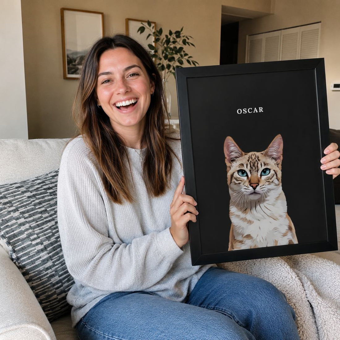 A woman sitting on a couch holds up a framed custom pet portrait featuring a detailed illustration of a cat named 'OSCAR' on a black background.